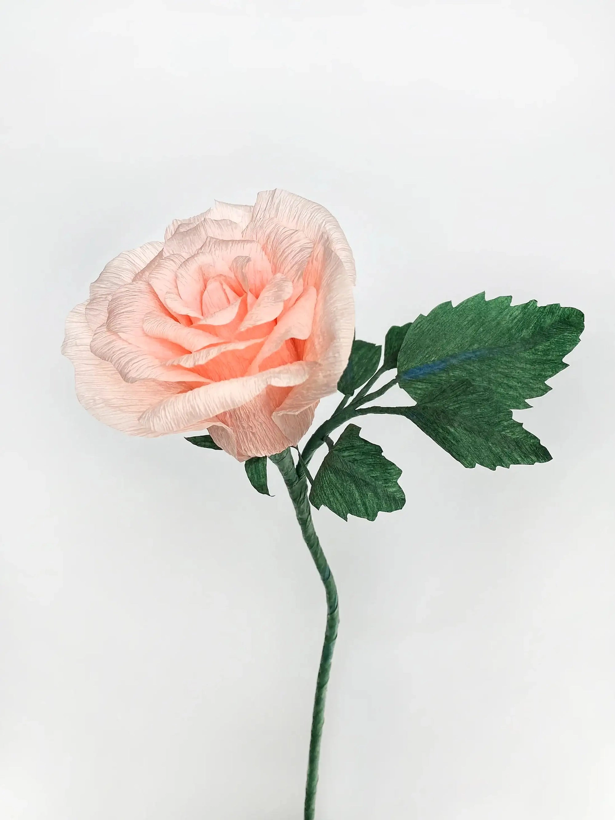 A handcrafted paper rose with delicately layered petals in soft shades of cantelope, curled and shaped to mimic the natural texture of a real rose. The flower is attached to a slender green stem with realistic-looking leaves made from emerald green crepe paper.